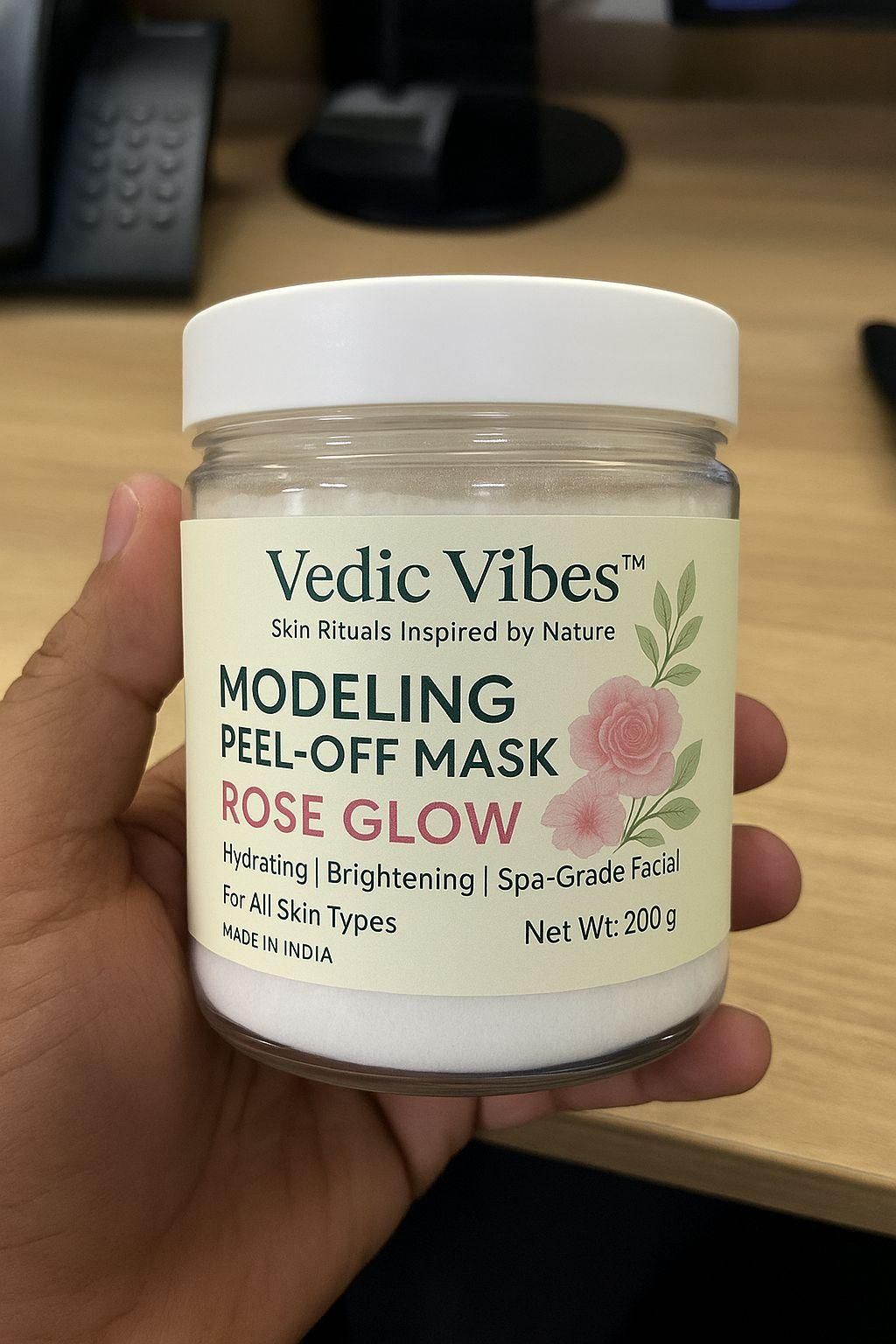Hand holding a jar of Vedic Vibes Modeling Peel-Off Mask with a blurred office background