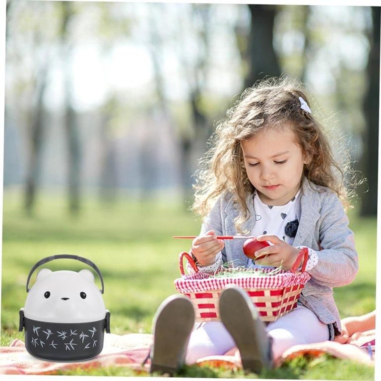 Child sitting on a blanket in a park with a black cartoon-style lunch bag and a red basket.