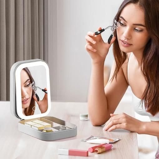 Woman using an eyelash curler in front of a compact mirror with LED lights.