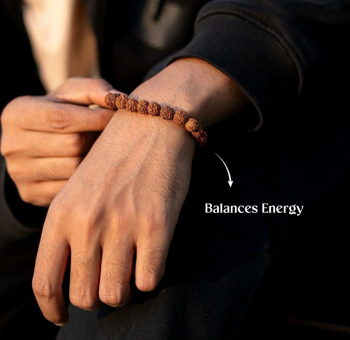 Hand wearing a rudraksha bracelet with text 'Balances Energy' on a dark background
